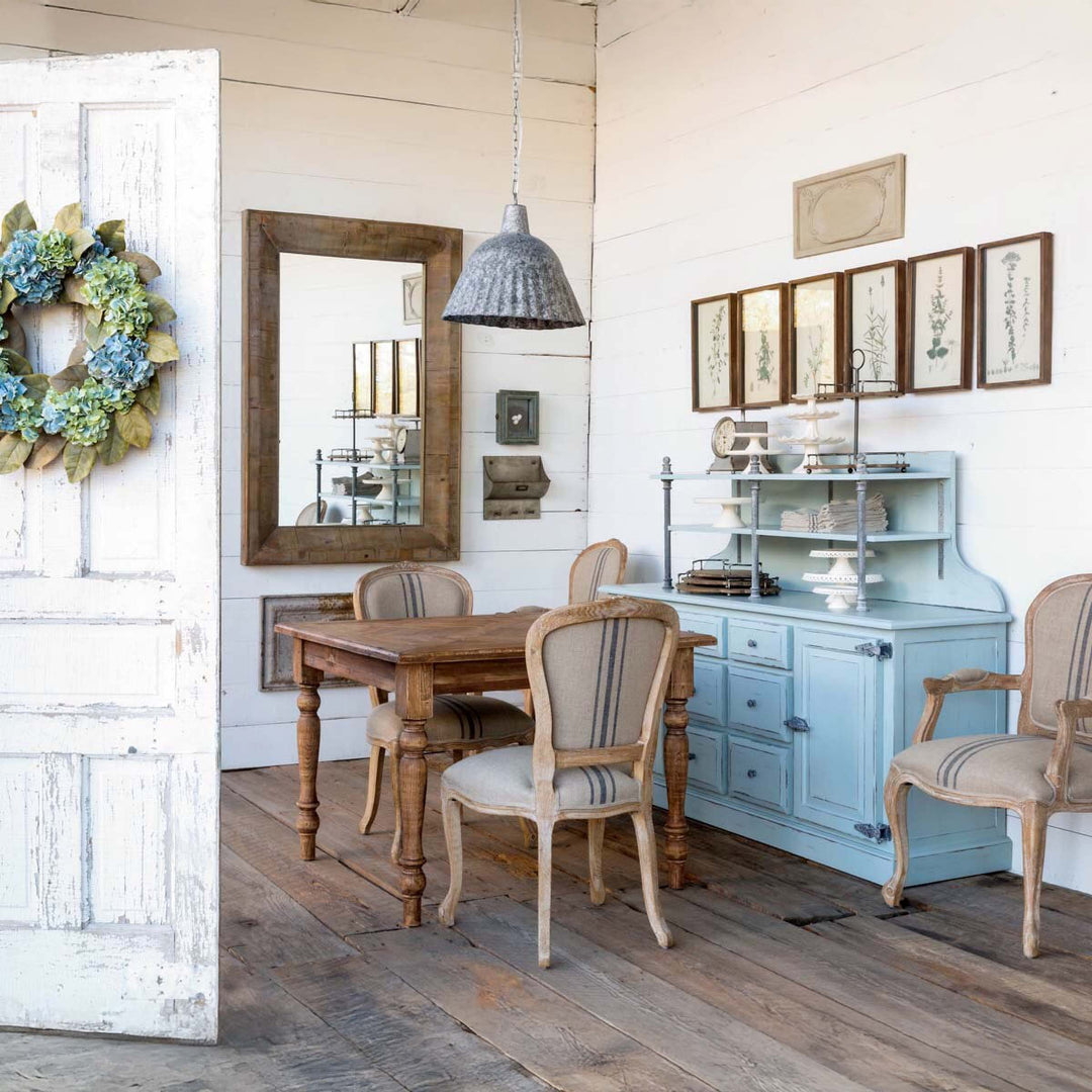Rustic vintage farmhouse dining room with wooden table, blue hutch, framed botanical prints, and a floral wreath on white door.