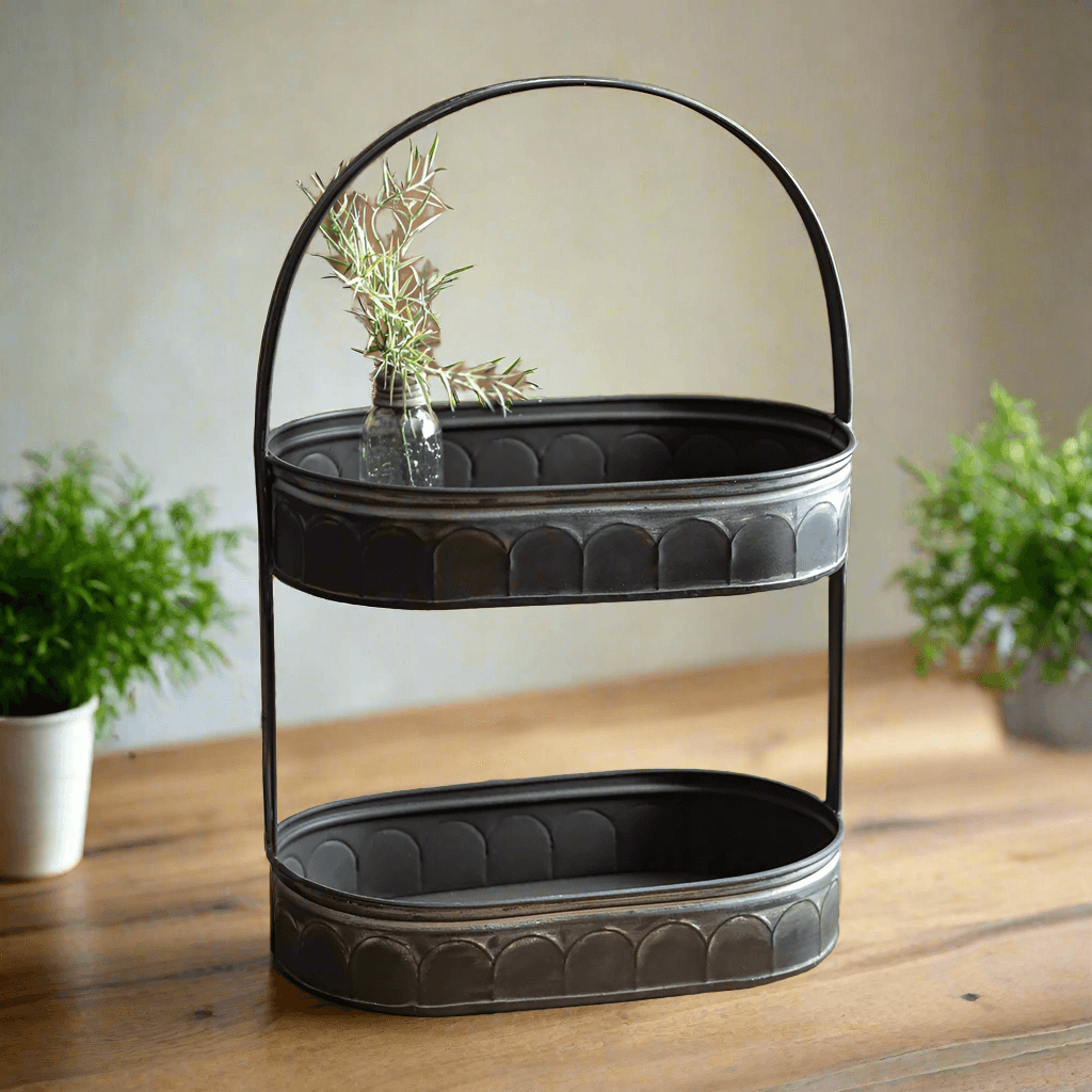 Vintage farmhouse black galvanized tiered tray organizer with arched handle, displaying a rustic tabletop with greenery accents.