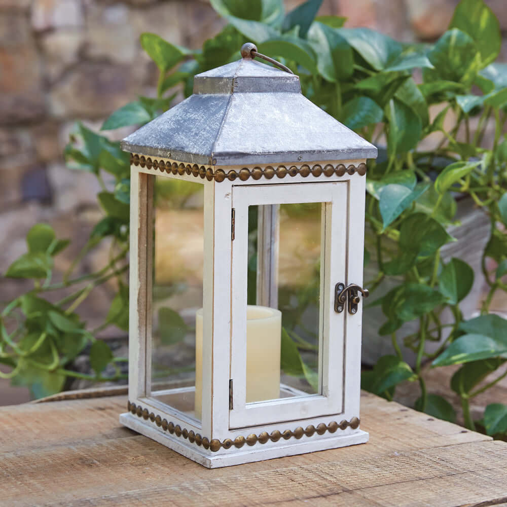 Rustic white farmhouse lantern with brass stud detail, featuring a warm candle inside on a wooden surface.