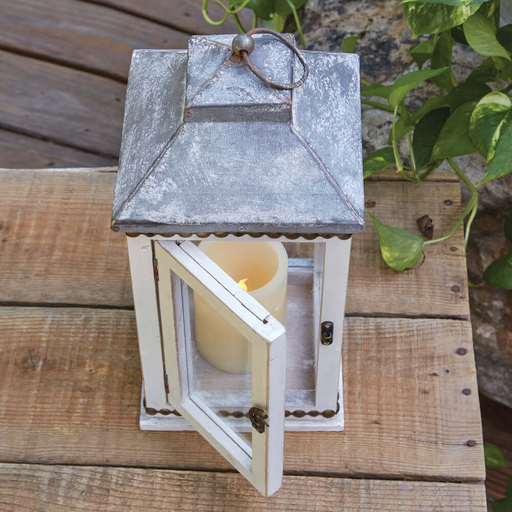 Rustic white farmhouse lantern with brass stud detail and candle inside, set on a wooden table.