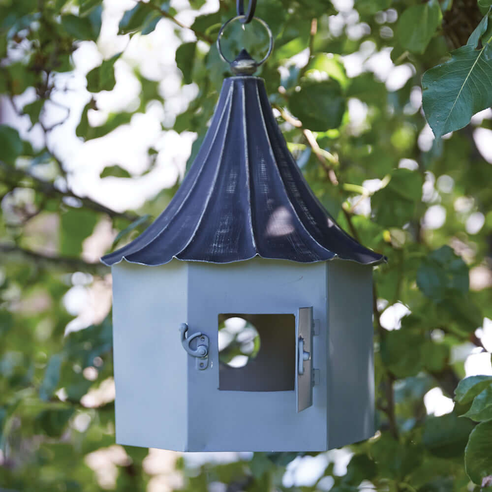 Victorian birdhouse with distressed metal roof hanging among green leaves, showcasing elegant craftsmanship.