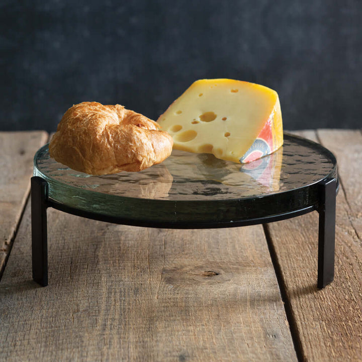 Cheese and bread on a glass stnd with a black metal stand on a wooden surface.