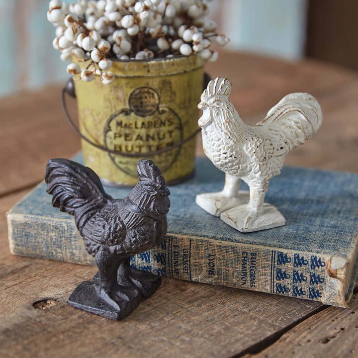 Set of 2 vintage cast iron rooster paperweights in brown and antique white, displayed on a book with a vintage tin.
