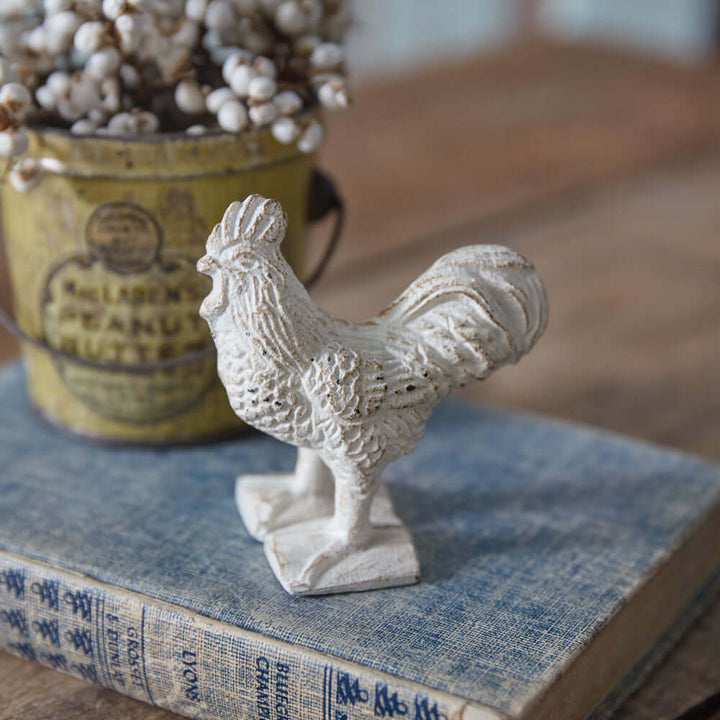 Vintage cast iron rooster paperweight in antique white, set on a blue book with farmhouse décor elements.