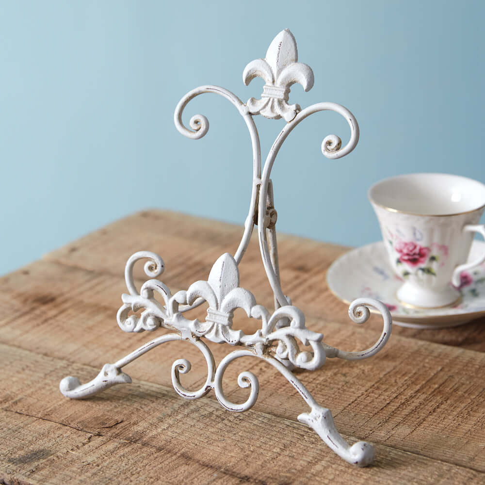 Vintage white metal easel stand with intricate scrollwork, perfect for displaying decorative items on a rustic table.