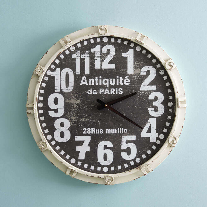 Vintage French country wall clock with bold white numbers on a weathered black face, set in a weathered metal frame against a pale background.