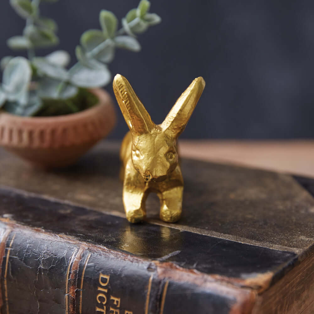Antique gold bunny figurine set on a vintage book with a potted plant in the background, adding nostalgic charm.