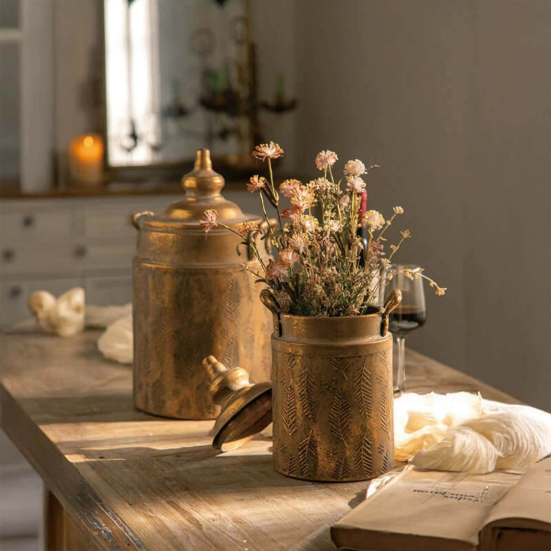 Antique brass embossed storage canisters with lids on rustic table, surrounded by flowers and soft light, evoking farmhouse charm.