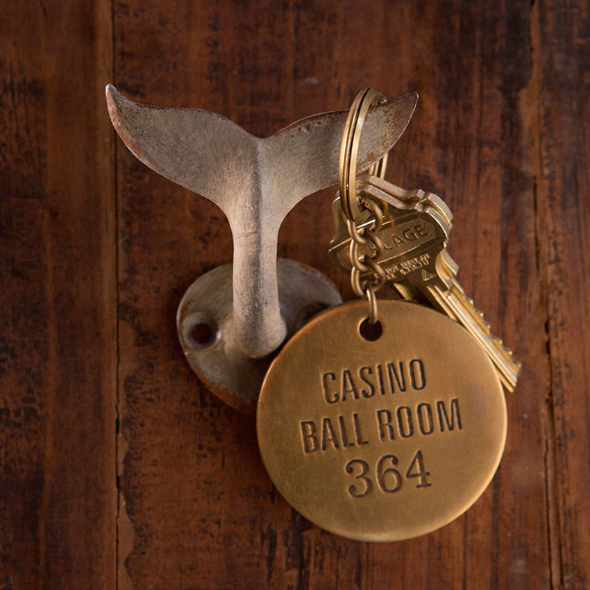 Distressed metal whale tail wall hook with brass key ring and casino room tag, ideal for coastal decor.