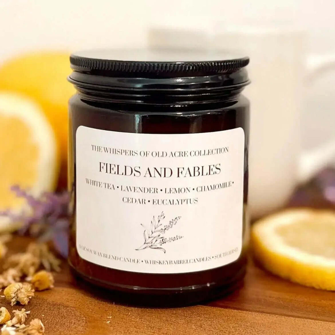 Fields and Fables Scented Candle: Chamomile, Lavender & Driftwood in dark jar, surrounded by lemons and flowers on wooden surface