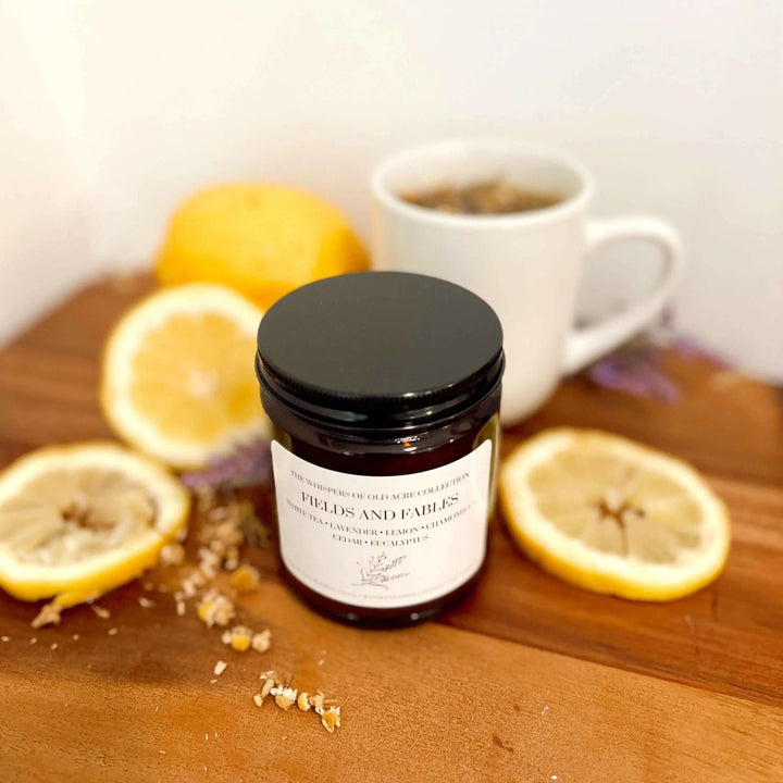 Fields and Fables Scented Candle: Chamomile, Lavender & Driftwood with lemons and tea on wooden table.