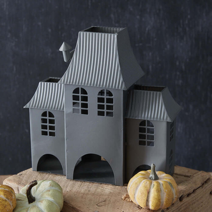 Metal tealight luminary shaped like a haunted house, with arched windows, dark finish, and small pumpkins, evokes vintage Halloween charm.