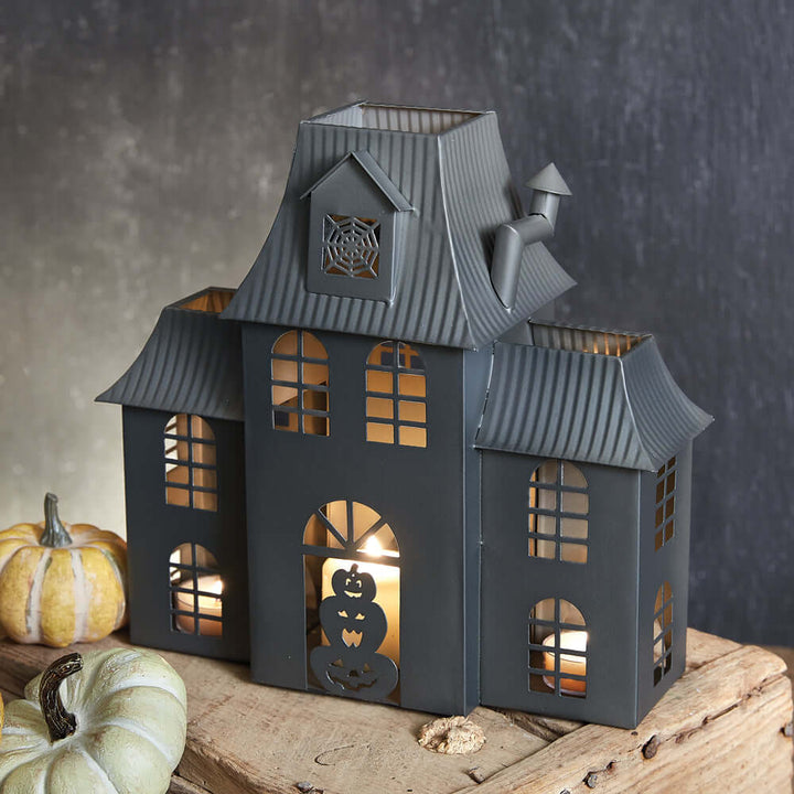 Haunted House Halloween metal tealight luminary with arched windows, capturing nostalgic fright and vintage tales; decorative pumpkins nearby.