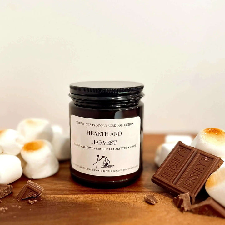 Hearth and Harvest Scented Candle: Marshmallow, Smoke & Eucalyptus with toasted marshmallows and chocolate on wood surface.