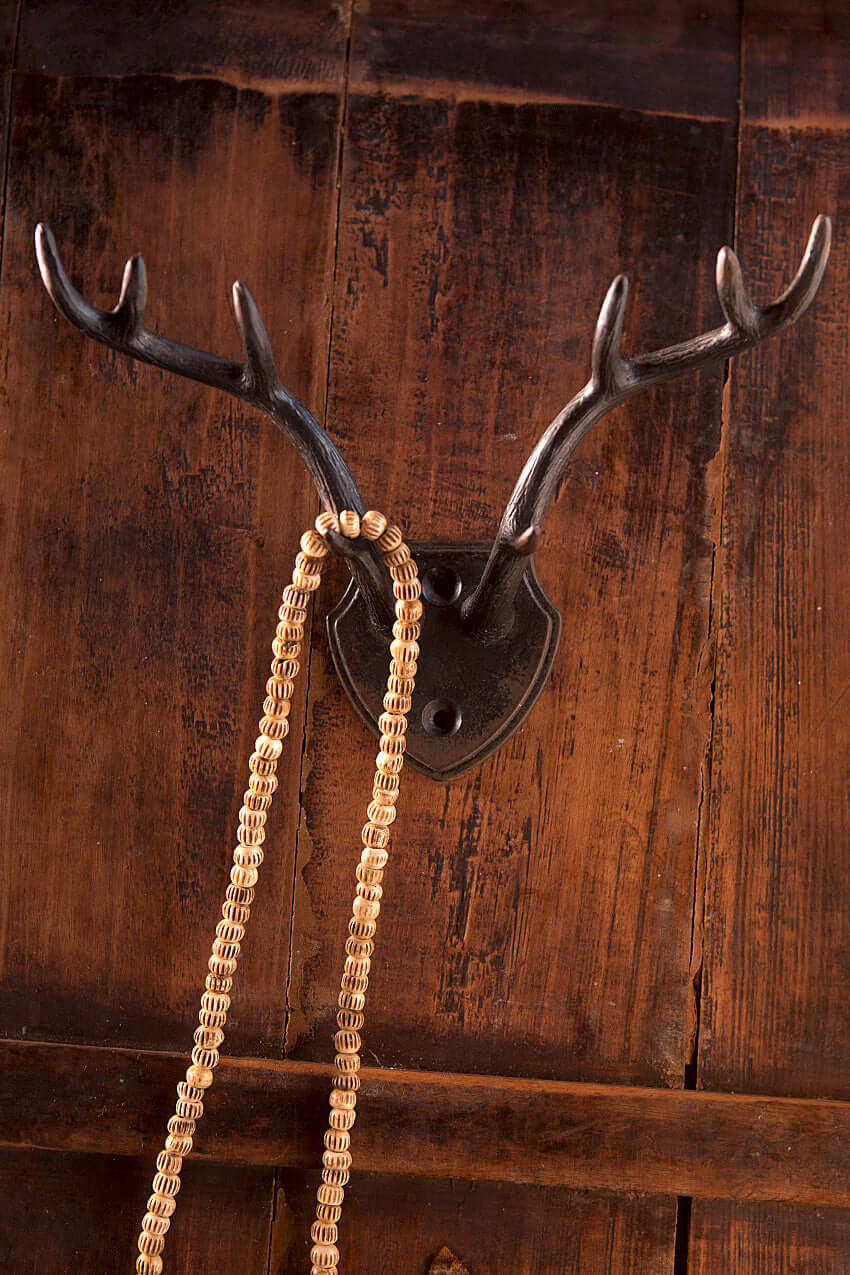 Rustic antler wall hook with shield backplate holding a braided necklace against a dark wooden background.