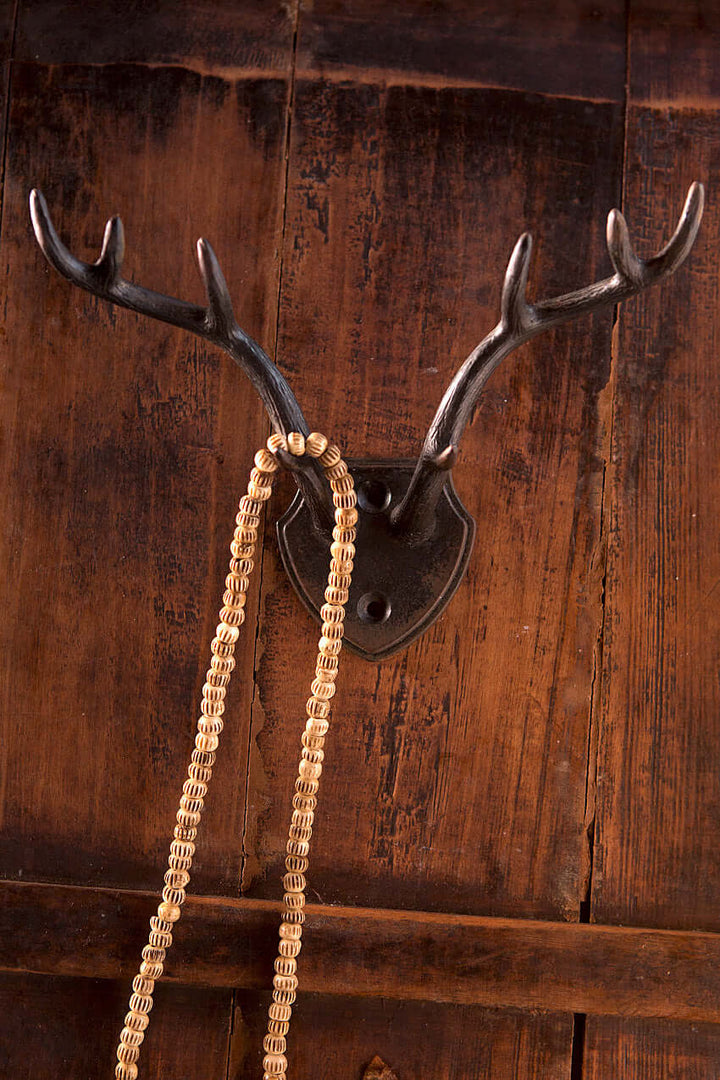 Rustic antler wall hook with shield backplate holding a braided necklace against a dark wooden background.