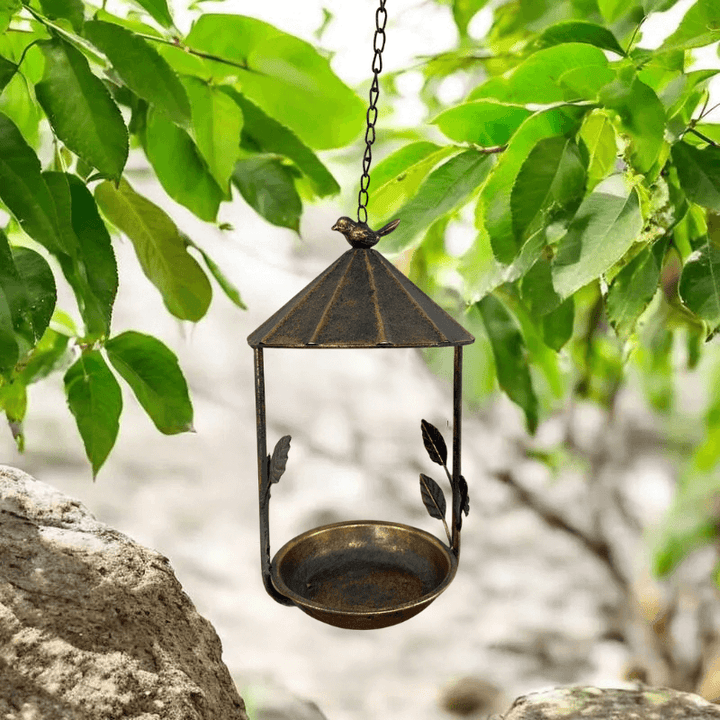 Rustic bronze hanging bird feeder with leaf and bird design set against green foliage background.