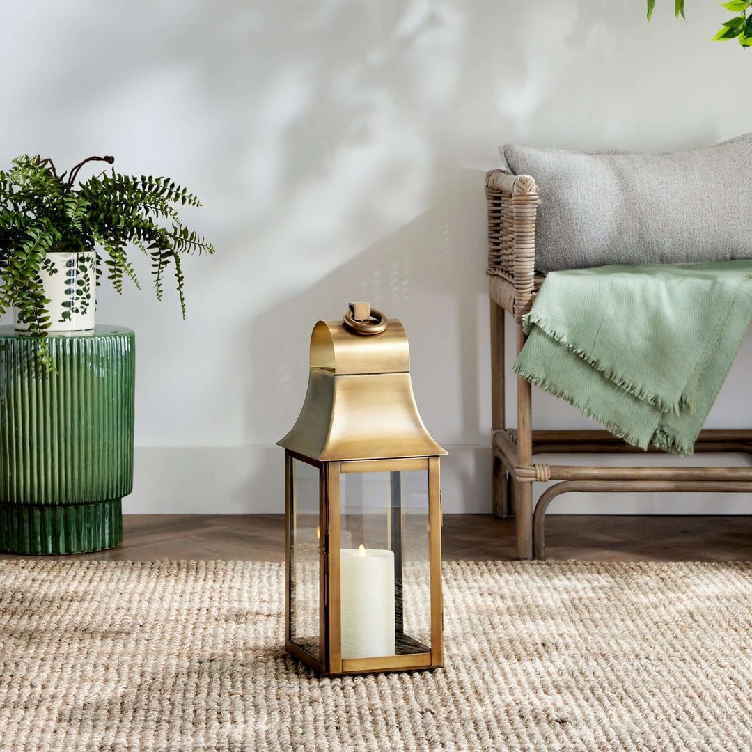 Antique brass candle lantern with hanging loop, exuding elegance and nostalgia, sits on a woven rug beside greenery and a cozy chair.