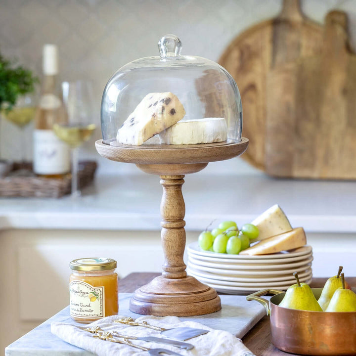 Tall rustic wood cake stand with glass dome displaying cheese in a cozy kitchen setting.