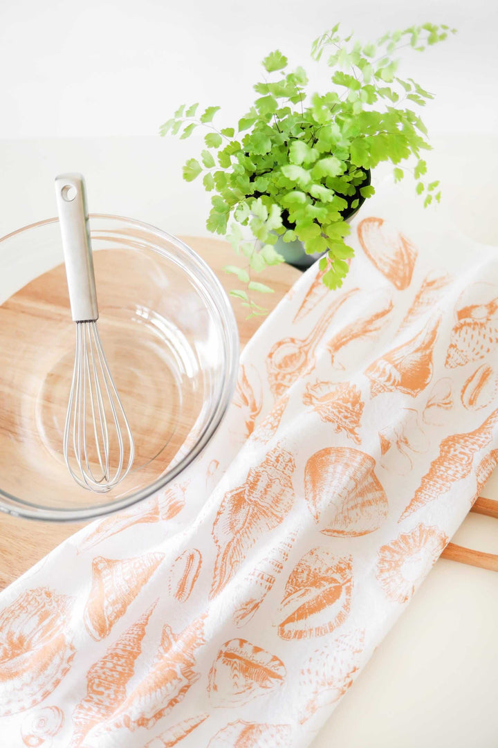 Vintage Coastal Seashell Tea Towel with watercolor-style seashells, displayed with a whisk and potted plant on a kitchen counter.