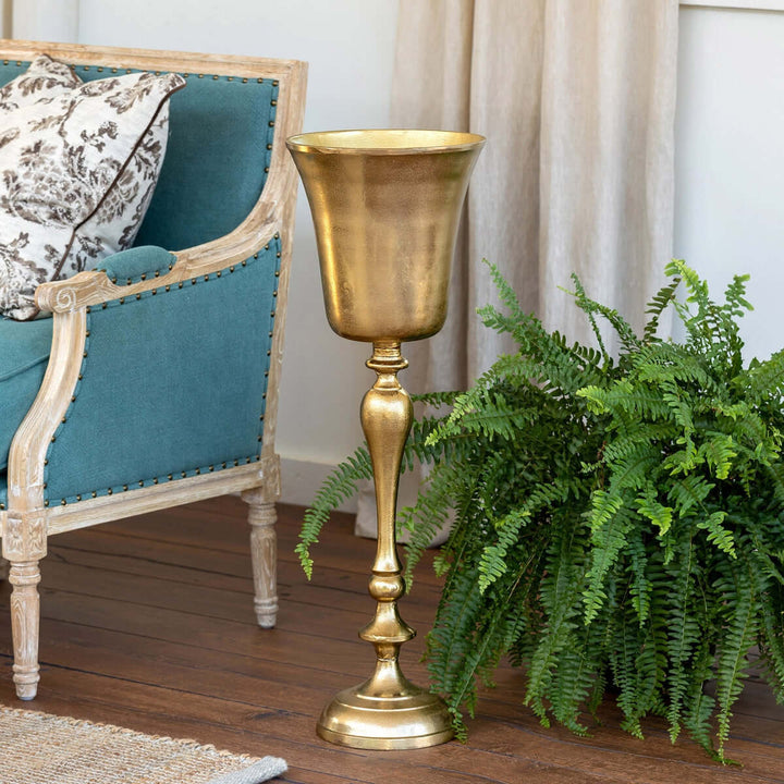 Vintage gold cast aluminum wine chiller with elegant design next to a classic armchair and lush greenery.