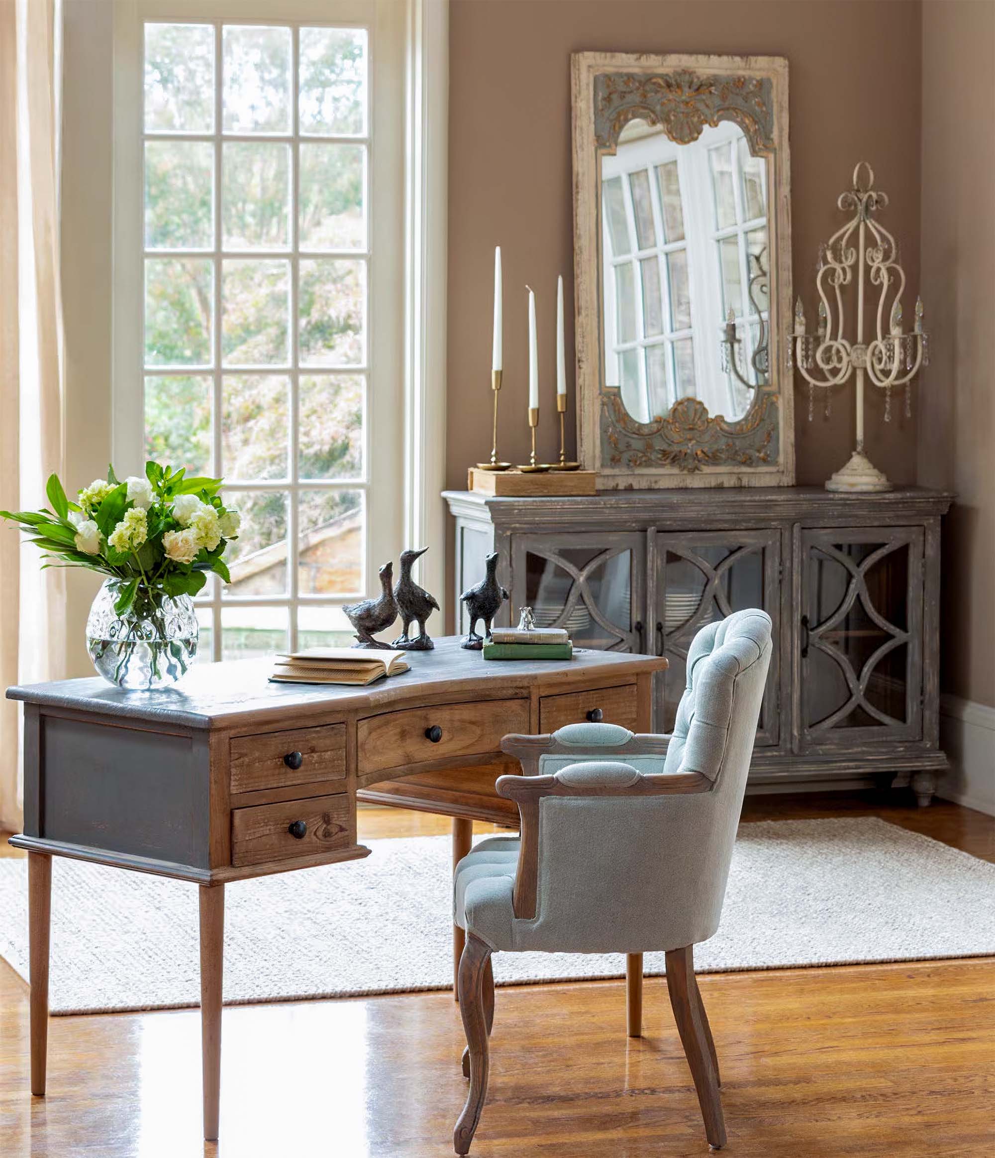 Elegant home office with wooden desk, plush chair, and decorative mirror reflecting light from large window.