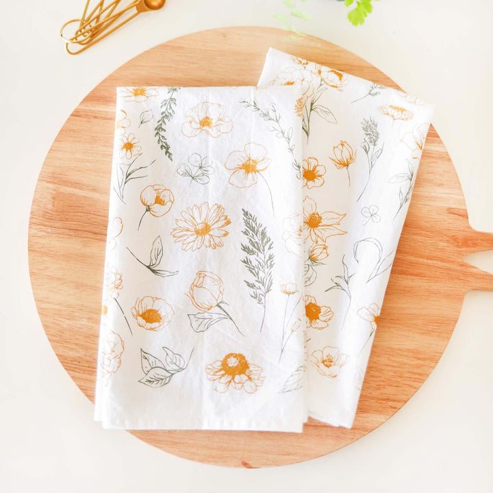 Vintage Botanical Flower Garden Tea Towel with watercolor-style blossoms in golden and green tones on a wooden surface.