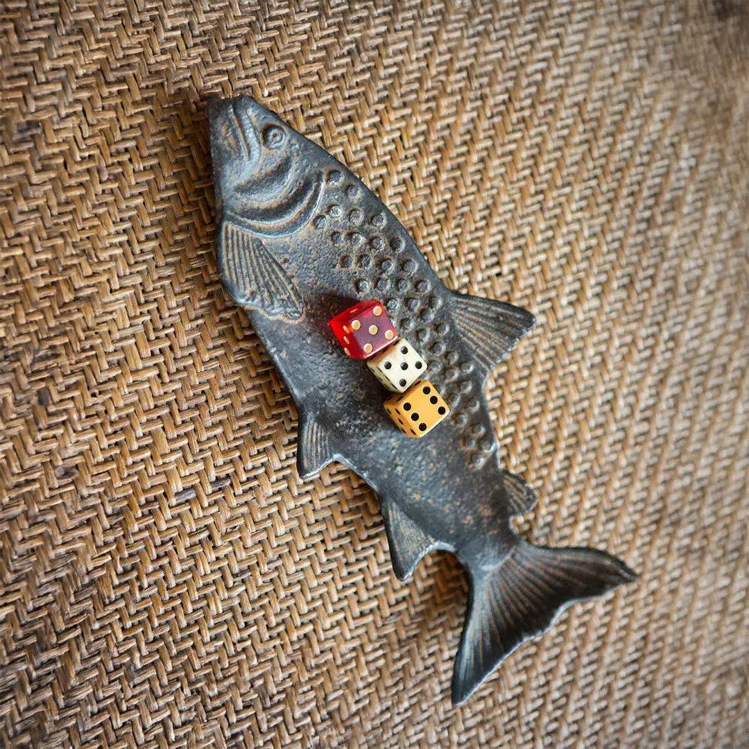 Vintage cast iron fish shaped tray with rustic patina on woven surface, featuring red and white dice for decorative appeal.