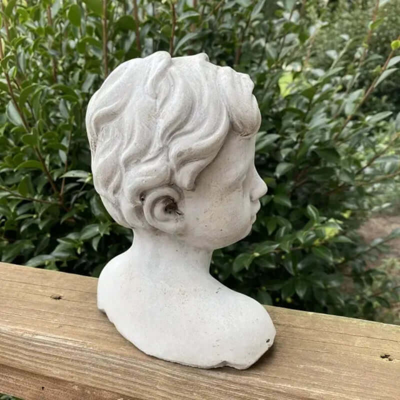 Side view of vintage cement boy bust garden statue with detailed curls, set against lush greenery on a wooden ledge.