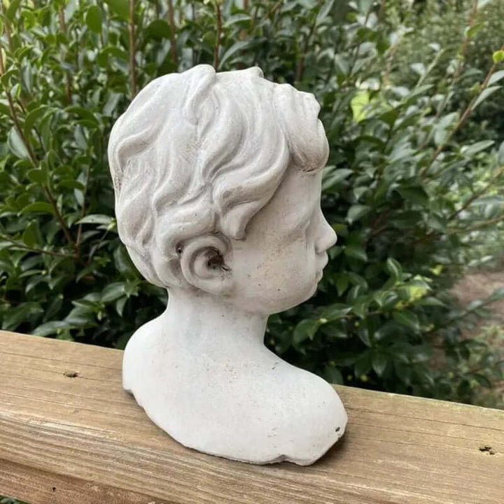 Side view of vintage cement boy bust garden statue with detailed curls, set against lush greenery on a wooden ledge.