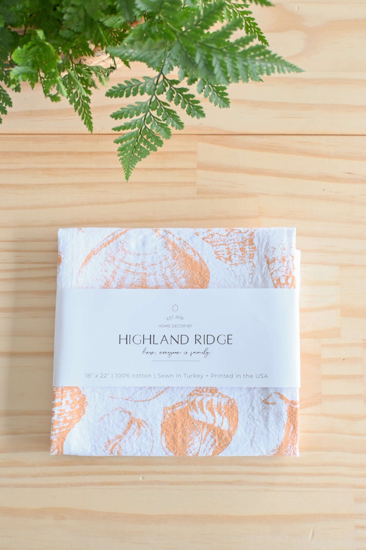 Vintage Coastal Seashell Tea Towel with delicate seashell design on a wooden table beneath a fern plant.