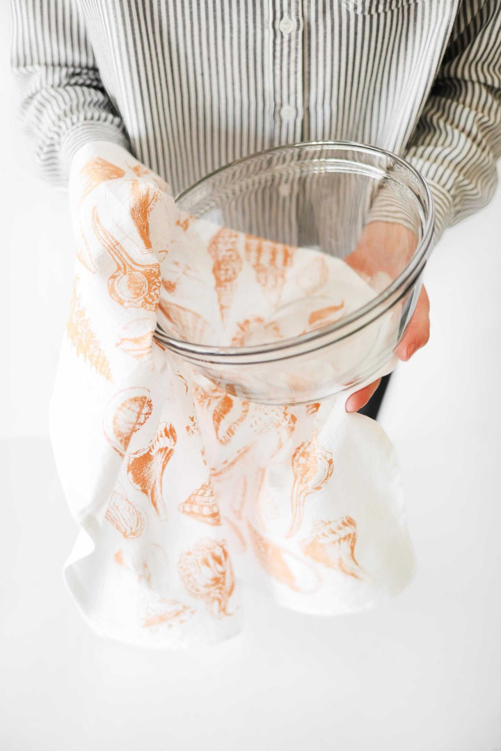 Person holding Vintage Coastal Seashell Tea Towel with glass bowl, featuring watercolor-style seashells for a seaside kitchen vibe.