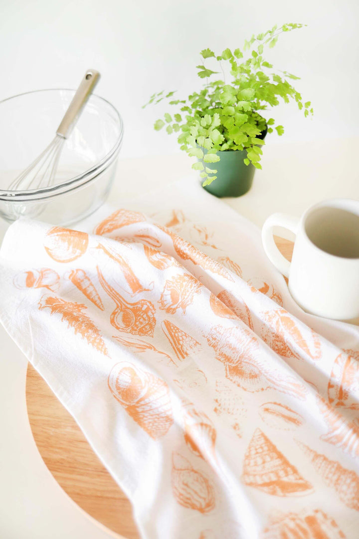 Vintage Coastal Seashell Tea Towel with seashell print, placed on kitchen counter beside a plant, bowl, and coffee cup.