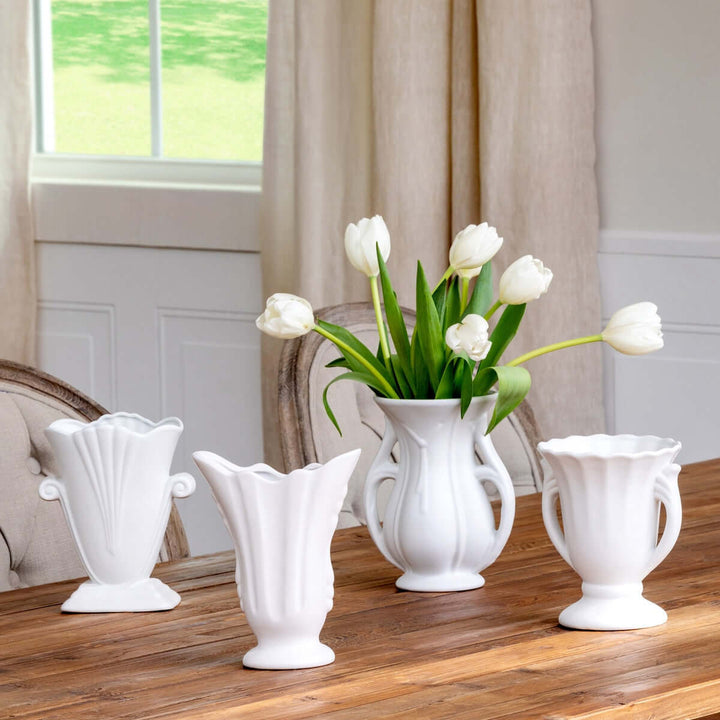 Set of 4 vintage-inspired white ceramic vases with tulips, featuring fluted necks and scalloped rims on a wooden table by a window.