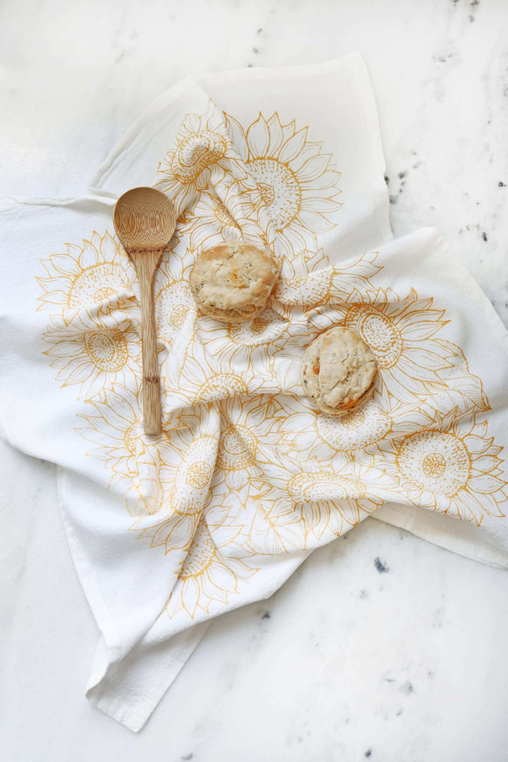Vintage Sunflower Tea Towel with golden watercolor sunflower design on cotton, displayed with wooden spoon and biscuits on marble countertop.