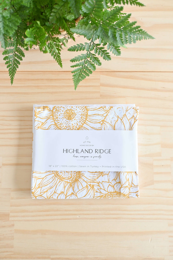 Vintage Sunflower Tea Towel with golden sunflower design on cotton, folded neatly on a wooden surface under green foliage.
