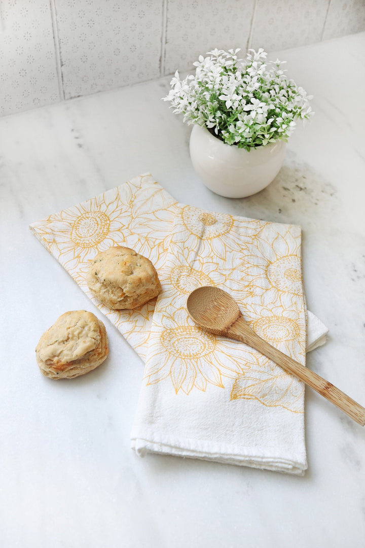 Vintage Sunflower Tea Towel with golden sunflower design on white cotton, placed on a kitchen counter with biscuits and a wooden spoon.