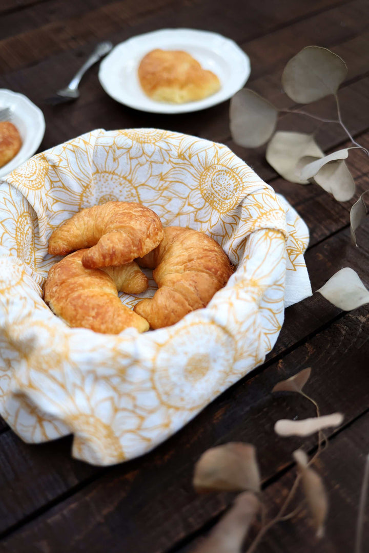 Fresh croissants wrapped in a Vintage Sunflower Tea Towel with golden sunflower prints, placed on a rustic wooden table.