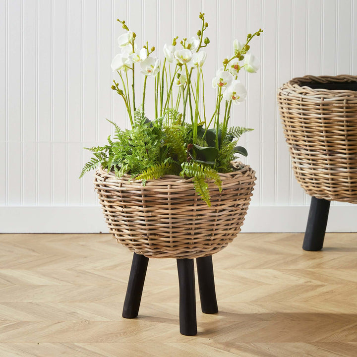 Woven rattan planter with black wood legs, featuring lush greenery and white orchids, enhancing natural decor.