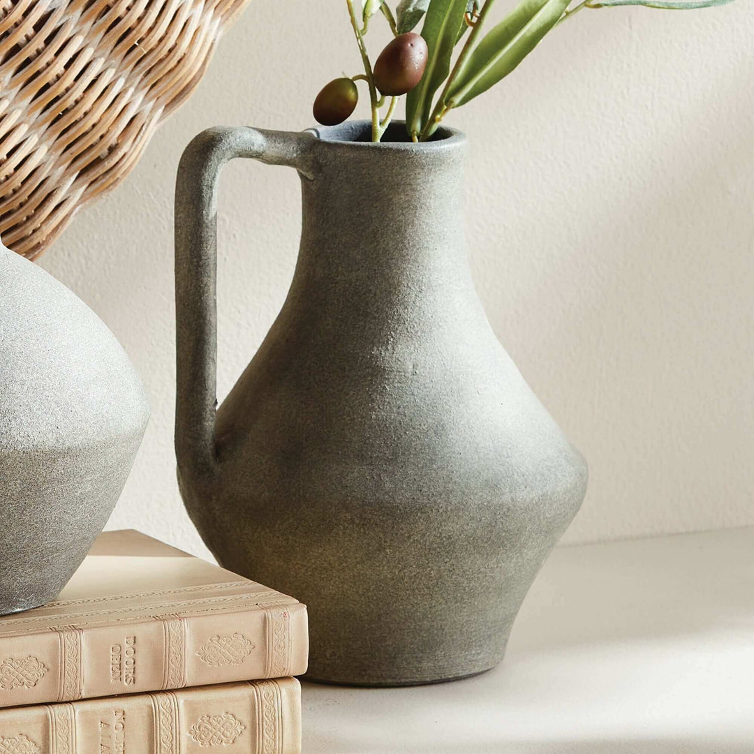 Weathered green-gray terracotta jug vase holding olive stems beside pale books and woven texture.