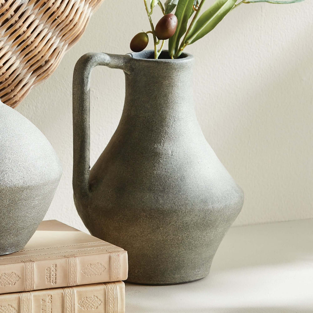 Weathered green-gray terracotta jug vase holding olive stems beside pale books and woven texture.