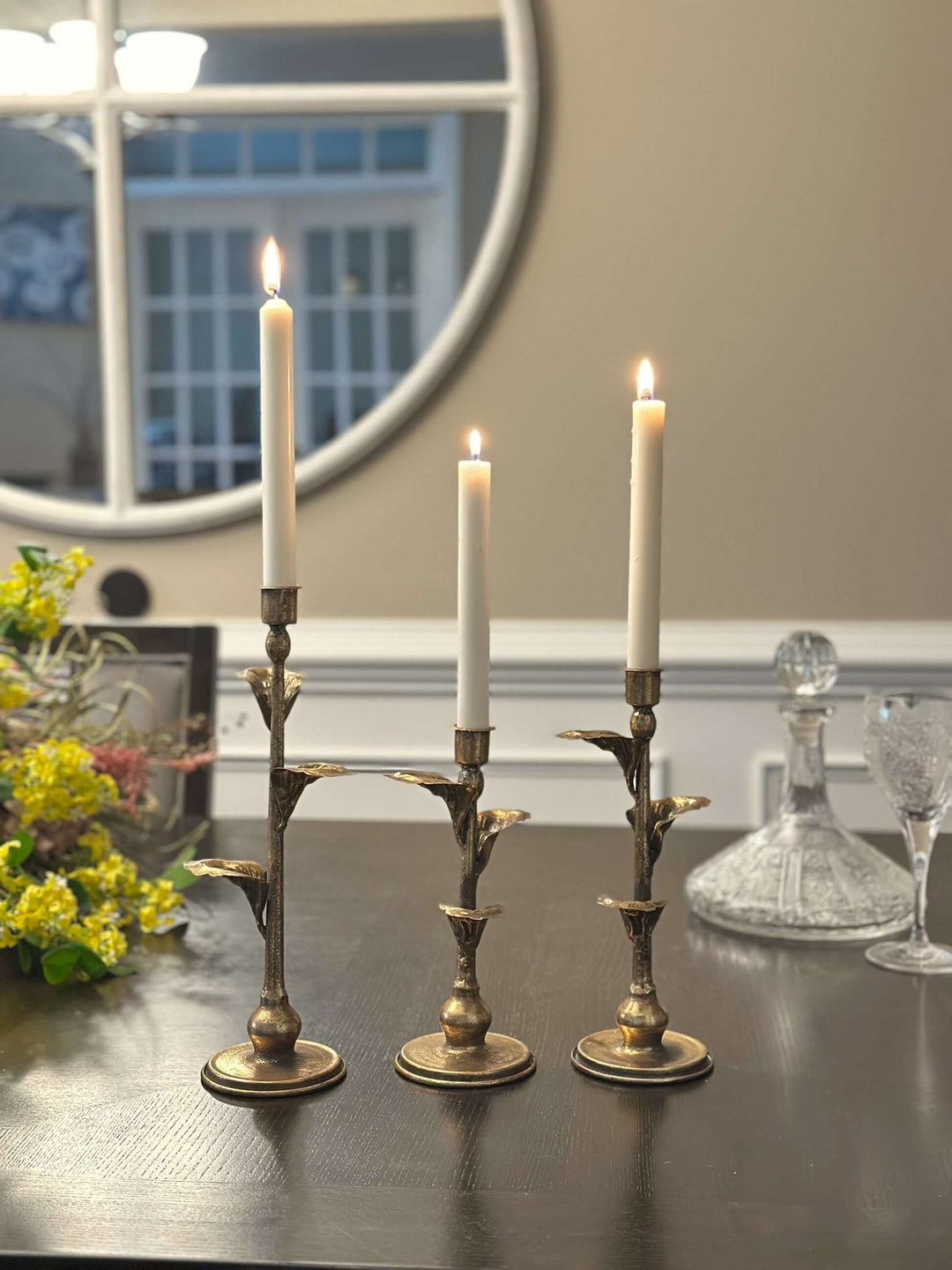 Three bronze-toned leaf candle holders with ivory tapers on a dark wooden dining table.