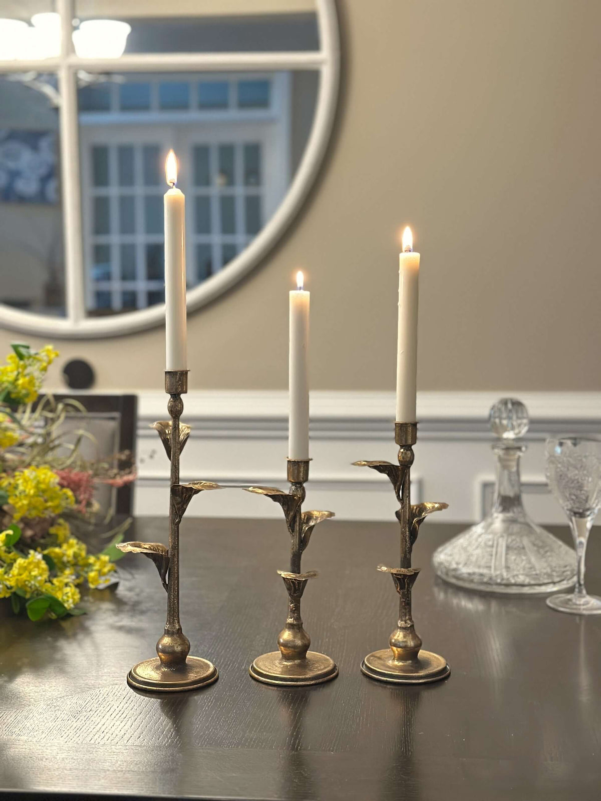 Three bronze-toned leaf candle holders with ivory tapers on a dark wooden dining table.