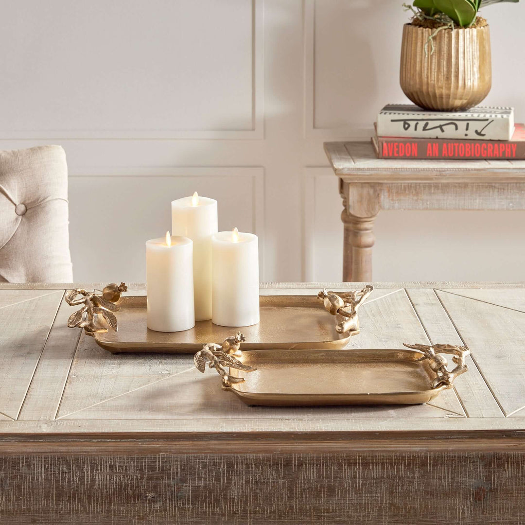 Two antique-gold pomegranate-handle trays with pillar candles on a wooden table