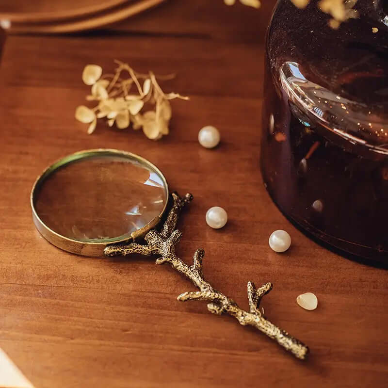 Gold-finished magnifying glass with a branching handle on a wooden table beside dried flowers and pearl-like beads
