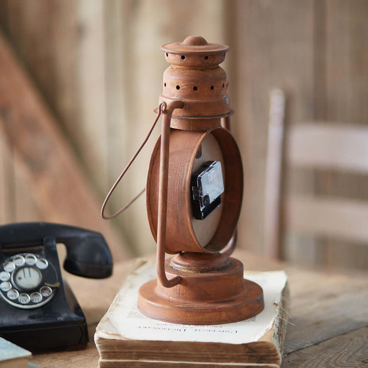 Vintage farmhouse lantern clock with rust finish resembling a kerosene lantern, evokes old homestead charm, on wooden table.