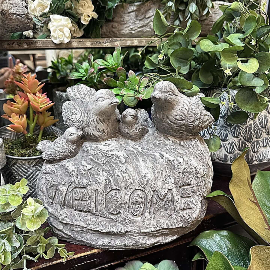 Vintage cement birds welcome sign with four songbirds on a stone base, surrounded by lush greenery.