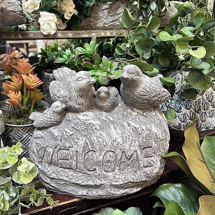 Vintage cement birds welcome sign with four songbirds on a stone base, surrounded by lush greenery.