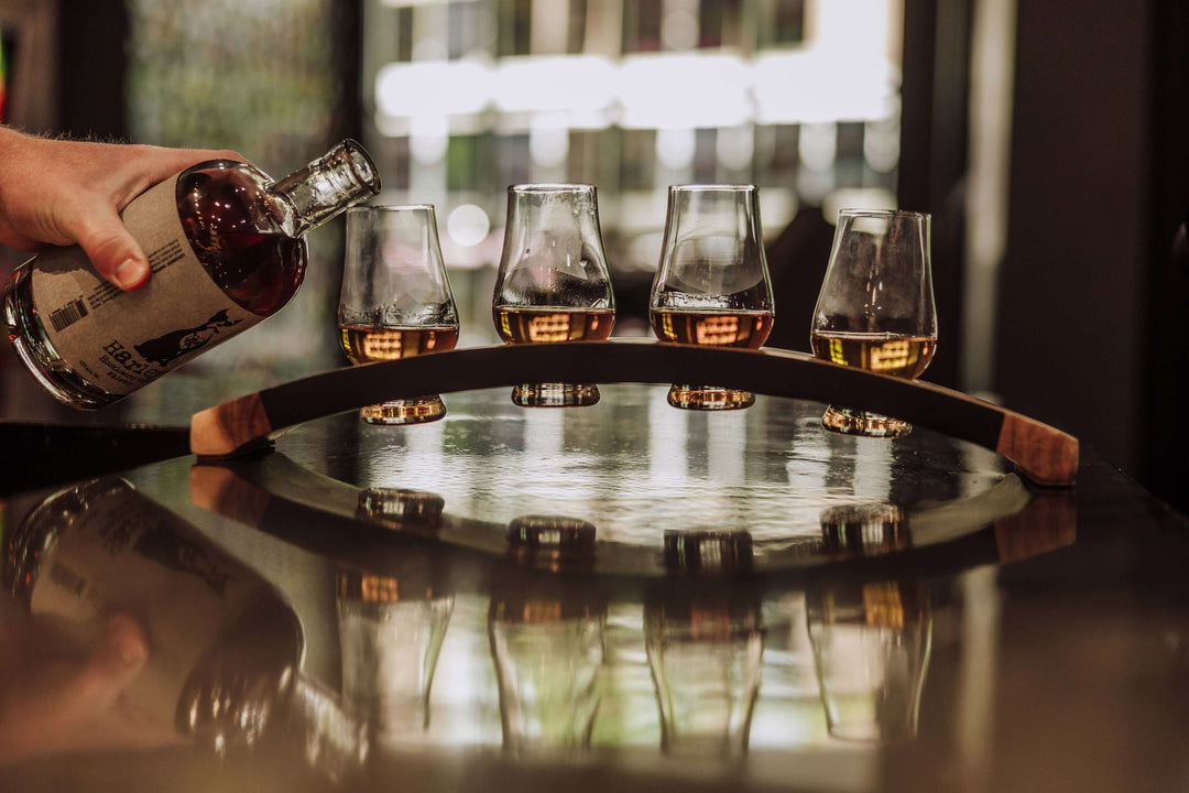 Person pouring whiskey from a bottle into four glasses on a wooden tray.