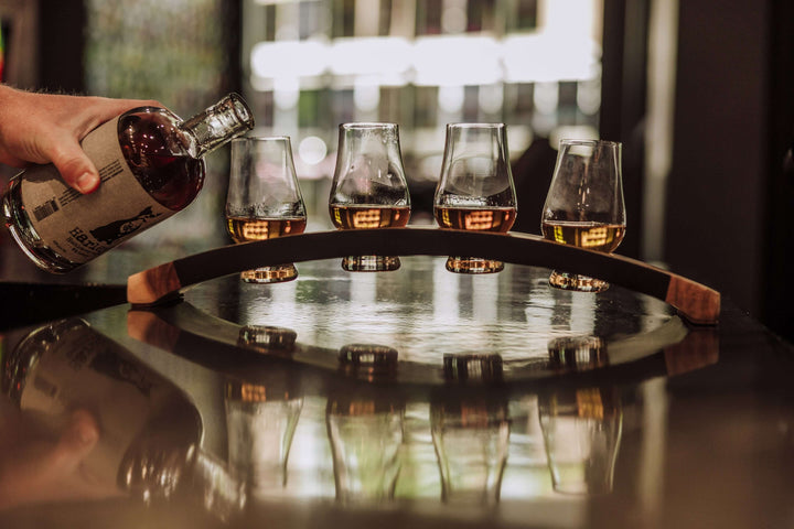 Person pouring whiskey from a bottle into four glasses on a wooden tray.